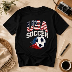 May include: Black t-shirt with a graphic design featuring "USA SOCCER" in white text and a soccer ball with red and blue streaks. The letters "USA" are designed with the American flag.