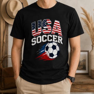 May include: Black t-shirt with "USA SOCCER" text and a soccer ball graphic. The text "USA" is designed with the American flag. The soccer ball has a blue, white, and red trail.