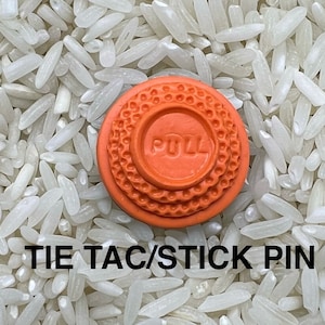 May include: An orange tie tac or stick pin with the word "PULL" in the center. The pin has a textured, circular design. The text "TIE TAC/STICK PIN" is at the bottom of the image. The background is white.