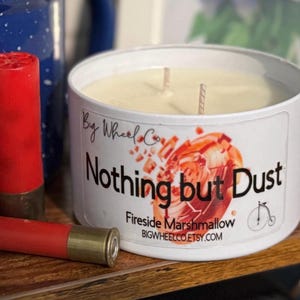 May include: A white candle in a tin with the text "Nothing but Dust" and "Fireside Marshmallow." Two red shotgun shells are next to the candle. The candle has two wicks and a graphic of a bicycle.