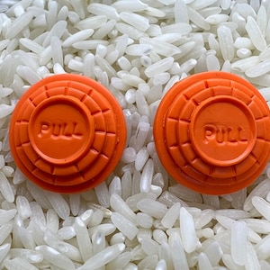 May include: Two orange clay targets with the word "PULL" embossed on the center. The targets are circular with concentric rings and are set against a background of white rice.