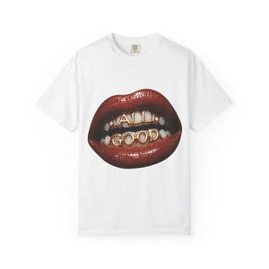 Può includere: Una t-shirt bianca a maniche corte con una grande stampa grafica di labbra rosse lucide. I denti sono decorati con grillz in metallo dorato che compongono il testo ALL GOOD in lettere maiuscole sulle file di denti superiori e inferiori.