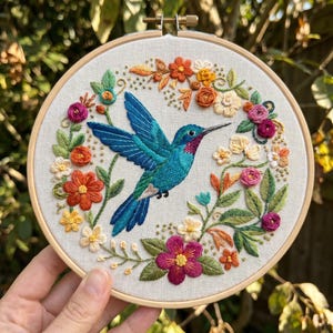 May include: A hand-stitched embroidery of a hummingbird in flight, encircled by flowers and leaves. The bird is teal with a red throat, set on cream fabric within a wooden hoop. The floral design includes orange, pink, and white flowers.