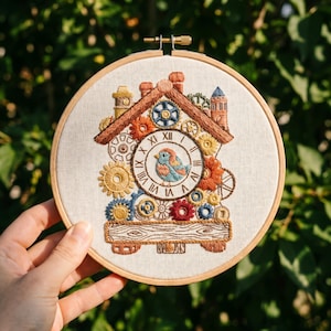 May include: A circular wooden embroidery hoop containing a fabric panel with a stitched cuckoo clock design. The clock face features Roman numerals and a central blue and orange bird. The design is surrounded by various colorful gear motifs.