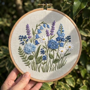 Può includere: Ricamo floreale su telaio di legno con fiori blu, viola e bianchi, tra cui campanule, ortensie e margherite. Le foglie sono verdi. Un'opera artigianale per l'arredamento della casa.