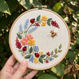 May include: A circular wooden embroidery hoop containing a cream-coloured fabric panel. The fabric features a hand-stitched floral wreath design with yellow, pink, and blue flowers, green leaves, red berries, three red ladybugs, and a bumblebee.