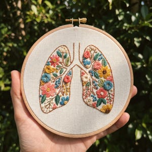 Peut inclure: Broderie représentant des poumons ornés de fleurs colorées et de feuilles vertes. Le motif est réalisé sur un tissu crème et encadré dans un cercle en bois. La broderie utilise diverses couleurs, dont le rose, le jaune et le bleu.