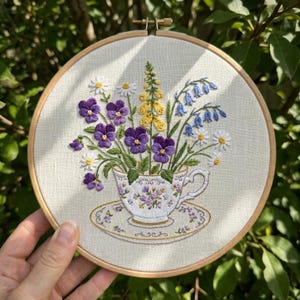 Op de afbeelding: Geborduurd bloemendesign in een houten ring. Het kunstwerk toont een theekopje dat overloopt van paarse viooltjes, gele vingerhoedskruid, blauwe klokjes en witte madeliefjes. De kop en schotel zijn wit met een bloemenpatroon.