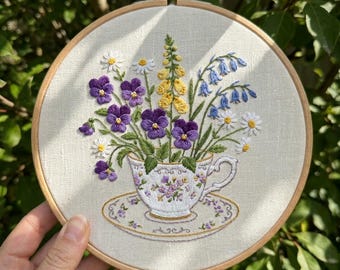 Peut inclure: Broderie florale dans un cercle en bois. L'œuvre représente une tasse à thé débordant de pensées violettes, de digitales jaunes, de campanules bleues et de marguerites blanches. La tasse et la soucoupe sont blanches avec un motif floral.