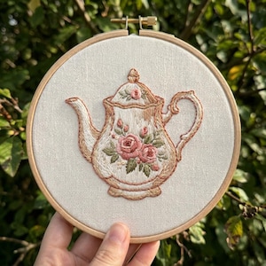 Può includere: Ricamo di un design di teiera in un telaio di legno. La teiera è color crema con finiture marroni e una composizione floreale di rose rosa e foglie verdi. Il ricamo è su uno sfondo di tessuto neutro.