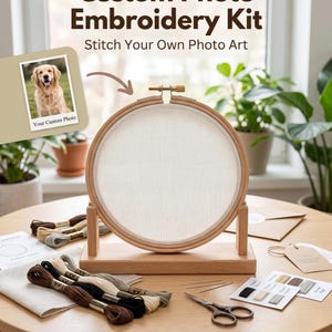 May include: A custom photo embroidery kit featuring a round wooden hoop on a stand, white fabric, assorted embroidery floss in brown, black, and cream, needles, scissors, a pattern guide, and a sample photo of a golden retriever dog.