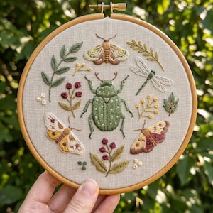 May include: A circular wooden embroidery hoop containing a light beige fabric panel. The fabric features hand-stitched embroidery of a large central green beetle surrounded by two moths, a dragonfly, and various botanical sprigs in green and red.