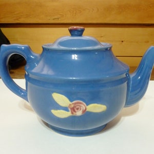 May include: A blue teapot with a single rose and leaves design. The teapot has a rounded body and a spout on the right side.