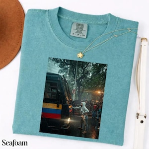 May include: A seafoam green short-sleeve t-shirt featuring a rectangular graphic print of a person standing next to a bus with yellow, blue, and red stripes on a city street. The shirt has a crew neckline and a Comfort Colors brand label inside.