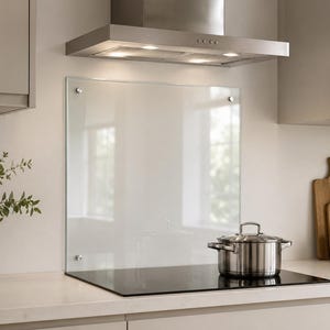 Könnte beinhalten: Eine rechteckige Küchenrückwand aus klarem Glas, die mit vier silberfarbenen, runden Befestigungselementen montiert ist. Die Platte ist vertikal hinter einem schwarzen Induktionskochfeld und einem Kochtopf aus Edelstahl angebracht.