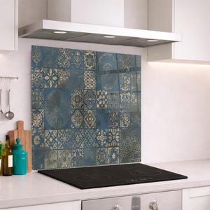 May include: A kitchen backsplash with a blue and gold tile pattern. The design features a variety of geometric and floral motifs. The backsplash is installed above a black stovetop and is surrounded by white cabinets and a range hood.