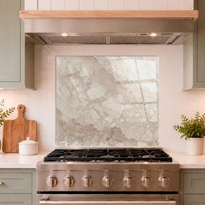 May include: A rectangular kitchen splashback panel featuring a glossy, light beige and grey marble stone pattern with intricate veining, installed on a white tiled wall behind a stainless steel gas range stove with six burners and silver knobs.