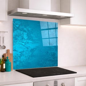 Op de afbeelding: Een rechthoekig spatbord voor de keuken met een levendig, getextureerd cyaanblauw oppervlak met een glanzende afwerking. Het paneel is verticaal geplaatst achter een zwarte inductiekookplaat, gemonteerd tegen een witte muur in een keuken.
