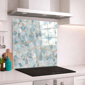 May include: A rectangular kitchen splashback panel featuring a light blue and white stone-like mosaic pattern with thin, copper-coloured veins running between the irregular shapes. The panel is positioned vertically behind a black induction hob.
