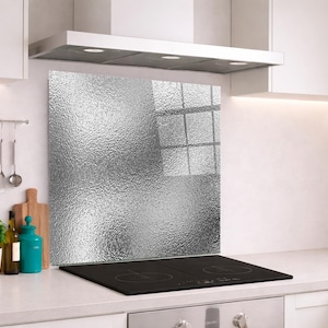 May include: A rectangular kitchen backsplash panel featuring a textured, metallic silver surface with a reflective, hammered finish. The panel is positioned vertically behind a black induction cooktop, mounted against a plain white wall.