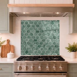 May include: A kitchen splashback panel featuring a repeating scalloped fish scale pattern in varying shades of sage green with white grout lines, positioned behind a stainless steel gas range cooker in a kitchen with sage green cabinetry.