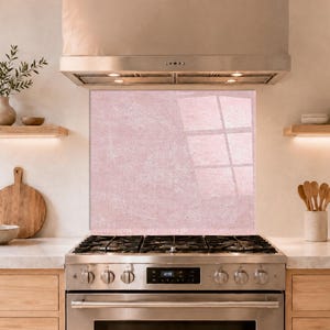May include: A rectangular kitchen splashback panel featuring a soft, dusty pink colour with a subtle, mottled stone-like texture. The panel is positioned vertically behind a stainless steel gas stove and range hood in a kitchen setting.