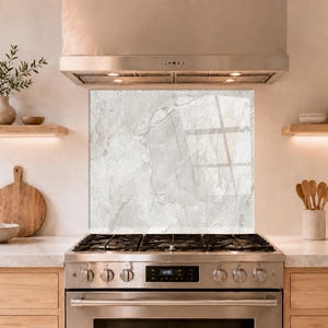 May include: A modern kitchen with a stainless steel gas range and a white marble backsplash. Wooden shelves hold decorative items, including a vase with greenery, a wooden cutting board, and a ceramic bowl. The range hood is stainless steel.