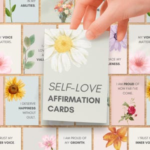 May include: A set of rectangular affirmation cards featuring floral illustrations and motivational text. The top card displays a large white daisy with a yellow center and the bold text SELF-LOVE AFFIRMATION CARDS in a dark gray sans-serif font.