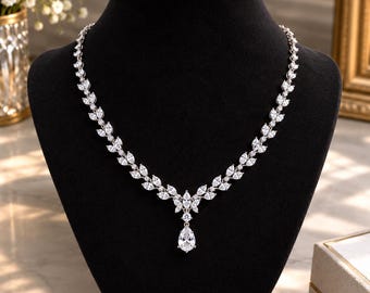 May include: A silver-toned necklace featuring a series of marquise-cut clear stones arranged in a leaf-like pattern, culminating in a central teardrop-shaped pendant. The necklace is displayed on a black velvet bust, showcasing the clear stones.