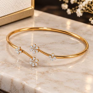 Op de afbeelding: Een open gouden armband met drie bloemvormige accenten. Elke bloem is ingelegd met kleine, heldere, fonkelende steentjes. De armband heeft een gladde, gepolijste afwerking, terwijl de secties naar de bloemen toe gedraaid zijn.