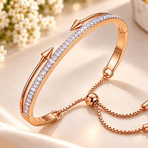 Puede incluir: Una pulsera en tono oro rosa con una hilera central de piedras claras brillantes, flanqueada por dos detalles en forma de flecha. Incluye una cadena ajustable con una cuenta deslizante y dos pequeños topes cilíndricos en los extremos.