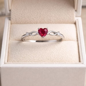 Puede incluir: Un anillo de tono plateado con una piedra preciosa roja central en forma de corazón. La banda muestra un diseño de infinito a ambos lados del corazón, acentuado con una pequeña piedra redonda transparente a cada lado, presentado en una caja color crema.