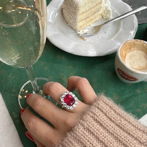 May include: A hand wearing a large, ornate ring with a vibrant red gemstone center and a halo of clear stones. The ring is set against a backdrop of a green tablecloth, a glass of champagne, and a slice of cake.