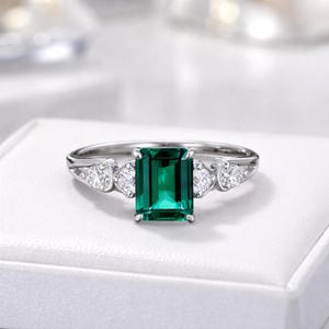 May include: A silver-toned ring featuring a central emerald-cut green gemstone flanked by two smaller, clear, round-cut stones on each side. The band is polished metal, and the setting holds the stones securely in a classic, symmetrical arrangement.