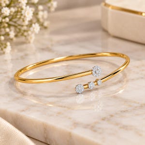 Puede incluir: Una pulsera abierta de oro con una banda pulida que termina en cuatro detalles circulares de diferentes tamaños, cada uno incrustado con pequeños diamantes blancos en pavé, creando una pieza de joyería delicada y refinada.