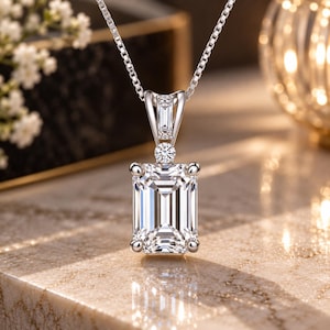 May include: A silver-toned necklace featuring a large emerald-cut clear gemstone pendant held by four prongs. Above the main stone sits a small round brilliant-cut stone and a baguette-cut stone set within the metal bail on a box chain.
