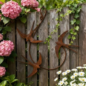 Könnte beinhalten: Drei rostige Metallschwalben sind an einem verwitterten Holzzaun befestigt. Die Vögel sind in einem dekorativen Muster angeordnet, wobei ein Vogel an einem gebogenen Metallstück befestigt ist. Rosa Hortensien und weiße Gänseblümchen umrahmen den Zaun.