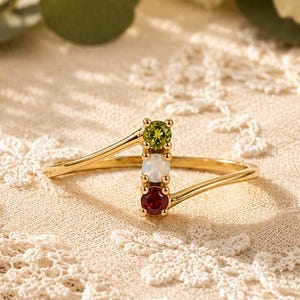 Puede incluir: Un anillo de oro con un diseño de banda cruzada y tres piedras preciosas de corte redondo engastadas verticalmente en el centro. Las piedras incluyen un peridoto verde arriba, un ópalo blanco en el medio y un rubí rojo en la parte inferior.