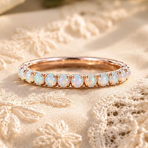 Op de afbeelding: Een roségouden ring met een doorlopende rij kleine, ronde, iriserende witte opaal-edelstenen in een zetting met pootjes. De ring is cirkelvormig en heeft een gepolijste metalen afwerking, waarbij het kleurenspel in elke opaalsteen zichtbaar is.