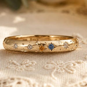 Könnte beinhalten: Ein goldener Bandring mit einer Reihe von sternförmigen Gravuren, die jeweils mit einem kleinen Edelstein besetzt sind. Die Steine umfassen klare, gelbe und blaue Varianten, die in einer Reihe auf der Oberfläche des polierten Metallrings angeordnet sind.