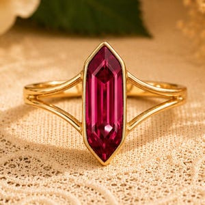 May include: A gold ring featuring a central, elongated hexagonal magenta gemstone. The band is crafted from thin, polished gold metal that splits into two delicate arms on each side, meeting at the base of the vibrant, faceted stone setting.