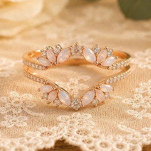 以下が含まれることがあります： バラの金色のリングガード、花の模様。リングは白いオパールのような石と小さな透明なクリスタルが特徴です。リングはクリーム色のレースのドイリーの上に置かれ、バラと緑のぼやけた背景があります。