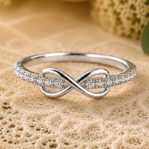 May include: A silver-colored ring with an infinity symbol design. The ring is encrusted with small, clear gemstones along the band and within the infinity symbol. The ring is set against a backdrop of lace and flowers.