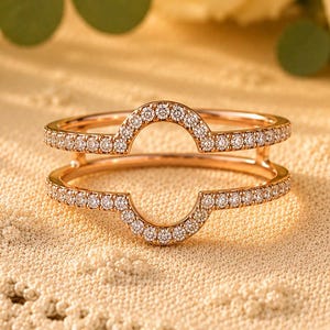 Puede incluir: Dos anillos de oro rosa con un diseño curvo, incrustados con pequeños diamantes brillantes. Los anillos están colocados sobre una superficie texturizada de color crema, con rosas amarillas en el fondo. Los anillos son una hermosa joya.