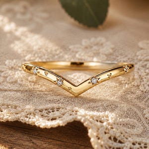May include: A gold V-shaped ring featuring a polished band with embedded small clear stones and engraved star-like patterns along the top surface. The ring has a chevron silhouette and a smooth, reflective metallic finish throughout the band.