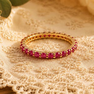 Könnte beinhalten: Ein goldener Ring mit einer durchgehenden Reihe leuchtend roter Edelsteine. Der Ring ist vor dem Hintergrund einer cremefarbenen Spitzendecke und unscharfen Rosen platziert, was eine elegante und romantische Ästhetik erzeugt.
