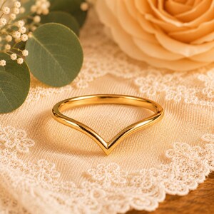 Pode incluir: Um anel de chevron dourado. O anel está sobre um tecido de renda creme, com um fundo desfocado de uma rosa cor de pêssego e vegetação. O design do anel é em forma de V simples.