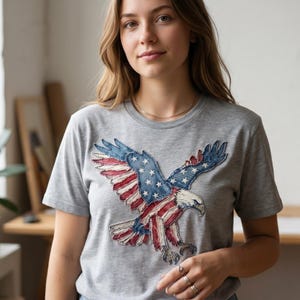 Puede incluir: Una camiseta gris con un diseño patriótico de un águila. Las alas del águila están adornadas con las estrellas y rayas de la bandera estadounidense. La camiseta está hecha de un material suave.