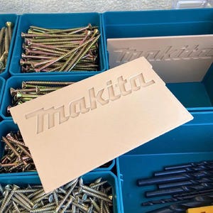 Puede incluir: Una caja organizadora de color turquesa llena de varios tornillos y brocas. Un divisor beige con el nombre de la marca "Makita" está colocado dentro de la caja. Los tornillos son dorados y plateados, y las brocas son negras.