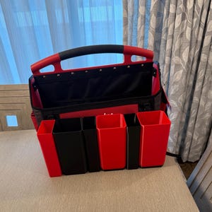 Puede incluir: Bolsa de herramientas roja y negra abierta con asa negra. La bolsa tiene múltiples bolsillos y compartimentos rojos y negros para organizar herramientas. La bolsa está sobre una superficie beige.
